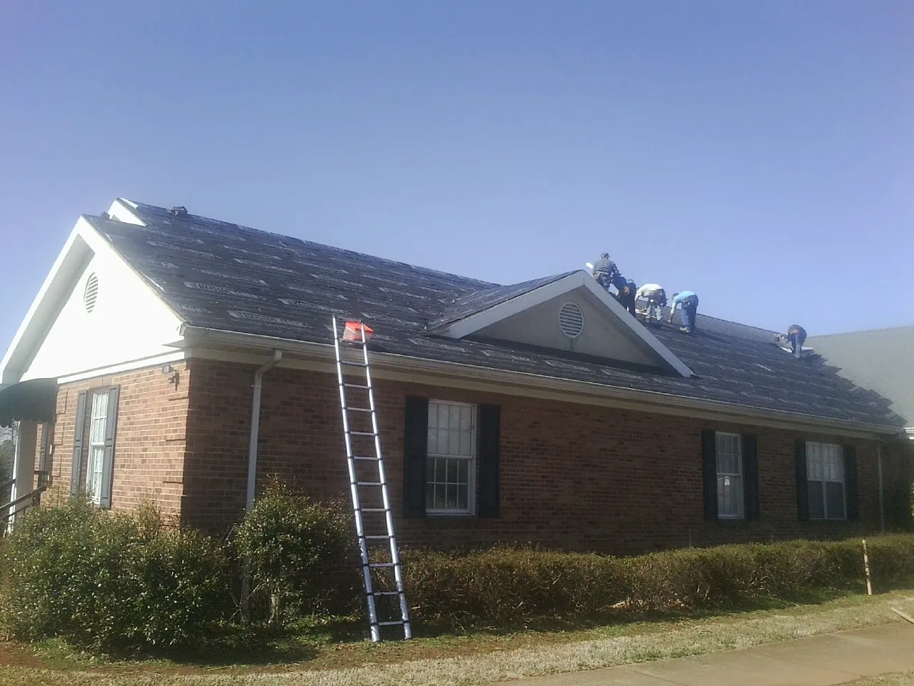 Crew tearing shingles off a brick ranch roof with a ladder set — Metal Roof Repair work by our crew in Jadwin