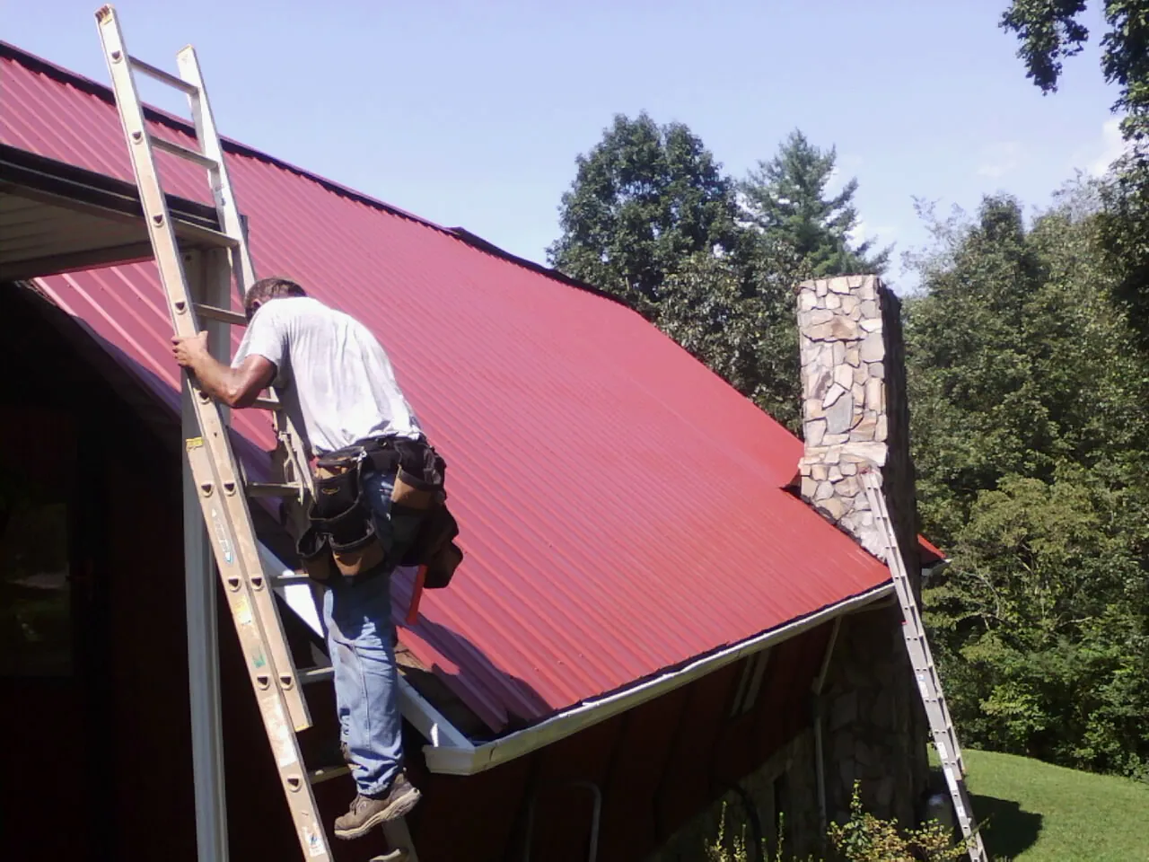 Roofer on a ladder beside a red metal roof and stone chimney — Soffit & Fascia Repair by Roofing Brothers in Jadwin